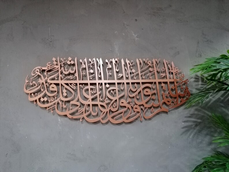 May include: A copper-colored metal wall art piece featuring intricate Arabic calligraphy. The design is in the shape of a crescent moon and features a repeating pattern of stylized letters.