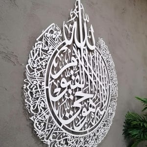 Op de afbeelding: Een groot, wit, cirkelvormig wandkunstwerk met ingewikkelde Arabische kalligrafie. Het ontwerp is lasergesneden met delicaat, onderling verbonden schrift dat aan de bovenkant een druppelvormig silhouet vormt.