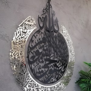 Op de afbeelding: Een cirkelvormig wandkunstobject met ingewikkelde Arabische kalligrafie. Het centrale gedeelte is matzwart, terwijl de buitenste ring een reflecterende zilveren afwerking heeft. Het ontwerp bestaat uit gelaagde metalen uitsparingen met gedetailleerd schrift, wat een ruimtelijk uiterlijk cre&euml;ert dat opvalt tegen het muuroppervlak.