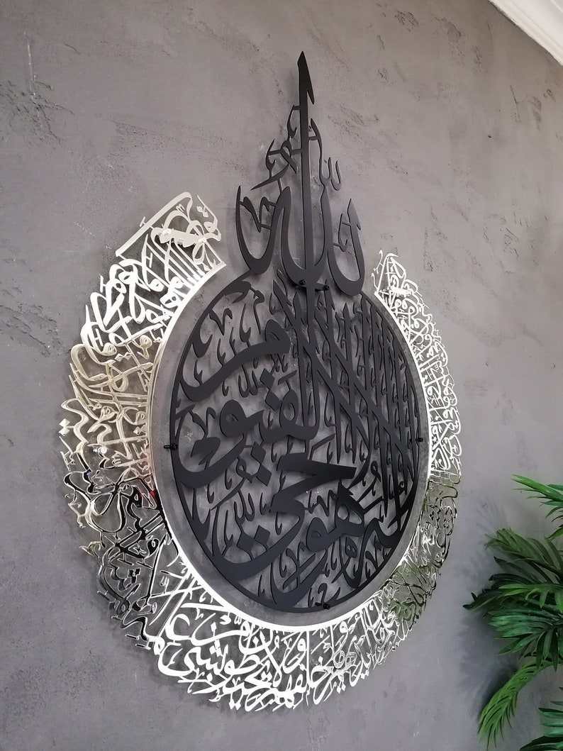 Op de afbeelding: Een cirkelvormig wandkunstobject met ingewikkelde Arabische kalligrafie. Het centrale gedeelte is matzwart, terwijl de buitenste ring een reflecterende zilveren afwerking heeft. Het ontwerp bestaat uit gelaagde metalen uitsparingen met gedetailleerd schrift, wat een ruimtelijk uiterlijk cre&euml;ert dat opvalt tegen het muuroppervlak.