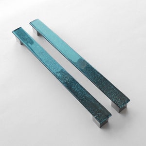 Op de afbeelding: Twee tealblauwe glazen kastdeurgrepen met een textuur en zilveren metalen bevestigingsplaten.
