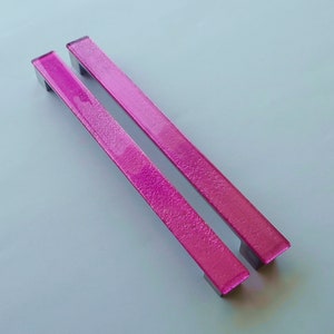 Peut inclure: Deux poignées de tiroir en verre rose avec une finition texturée. Les poignées sont rectangulaires et ont un bord légèrement arrondi.