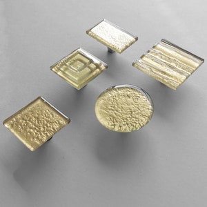 Może przedstawiać: Zestaw pięciu dekoracyjnych gałek do szafek z pozłacanymi, teksturowanymi szklanymi powierzchniami. Gałki mają różne kształty: prostokątny, rombowy i okrągły, każda z podstawą z posrebrzanego metalu. Szkło ma błyszczący, artystyczny wygląd.