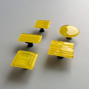 May include: Five yellow glass cabinet knobs with black metal bases. The knobs are in various shapes, including square, rectangular, and round.