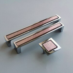 Peut inclure: Trois poignées de tiroir argentées avec un insert en verre texturé rose. Les poignées sont de tailles différentes, la plus petite étant de forme carrée.