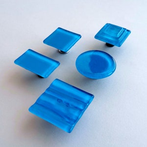 Peut inclure: Cinq boutons de cabinet en verre bleu de formes différentes, notamment carrées, rondes et rectangulaires. Les boutons ont une finition en verre dépoli et sont montés sur des bases en métal argenté.