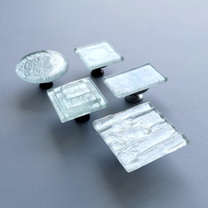 Peut inclure: Cinq boutons de cabinet en verre givré avec quincaillerie en métal argenté. Les boutons sont de formes variées, notamment rondes, carrées et rectangulaires.