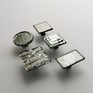 May include: Five silver and black glass cabinet knobs with different patterns. The knobs are square and round in shape.