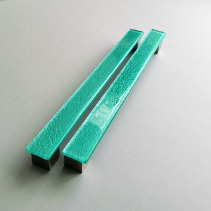 May include: Two rectangular, turquoise glass cabinet pulls with a textured surface. The pulls are mounted on square, dark metal bases. The pulls are displayed on a light gray background.