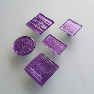 Op de afbeelding: Vijf paarse glazen kastknoppen in verschillende vormen, waaronder een cirkel, een rechthoek, een vierkant en twee diamanten.
