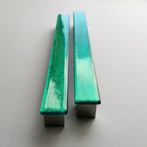 Op de afbeelding: Twee groene glazen kastgrepen met een metalen afwerking. De grepen zijn rechthoekig en hebben een getextureerd oppervlak.