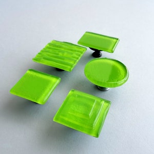 Op de afbeelding: Vijf limoengroene glazen kastknoppen met verschillende vormen en texturen. De knoppen zijn allemaal bevestigd aan zilveren metalen bases.