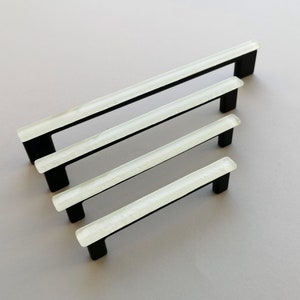 Pode incluir: Quatro puxadores de armário de vidro branco de diferentes tamanhos com ferragens de metal preto. Os puxadores estão dispostos em forma de escada, com o puxador maior na parte superior e o menor na parte inferior.