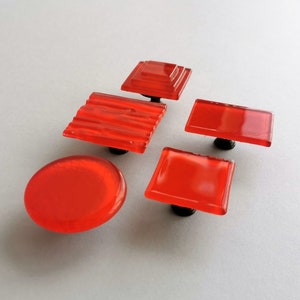 Pode incluir: Cinco botões de armário de vidro vermelho com diferentes formas e texturas. Os botões são todos presos a bases de metal preto.