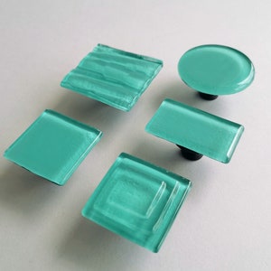 May include: Five turquoise glass cabinet knobs in various shapes: square, rectangular, and oval. Each knob has a black base. The glass has a textured, translucent appearance, adding a decorative touch to furniture.