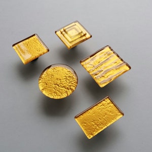 Może przedstawiać: Pięć dekoracyjnych gałek do szafek o różnych kształtach: kwadratowych, rombowych i okrągłych. Gałki mają wzór ze złotych liści zamknięty w przezroczystym szkle z bursztynowymi krawędziami. Okucia są zamontowane na szarej powierzchni.