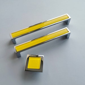 Może przedstawiać: Dwa chromowane uchwyty do szafek z wkładkami ze szkła żółtego i jeden mniejszy chromowany gałka z wkładką ze szkła żółtego.