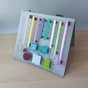 Peut inclure: Un présentoir en métal avec le texte "CITY GLASS STUDIO" présente divers échantillons de verre colorés. Les échantillons comprennent des pièces rectangulaires et carrées dans des tons de bleu, vert et rose, ainsi que des tiges de verre transparentes et texturées.