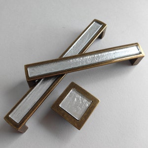 May include: Two silver and brass cabinet pulls with a square knob. The pulls are rectangular with a silver inlay and a brass frame.