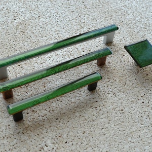 Può includere: Un set di ferramenta per mobili con tre maniglie rettangolari e un pomello quadrato. Le maniglie hanno un tubo di vetro trasparente riempito di dettagli verdi e bianchi, con basi argentate, marroni e nere. Il pomello quadrato è verde.