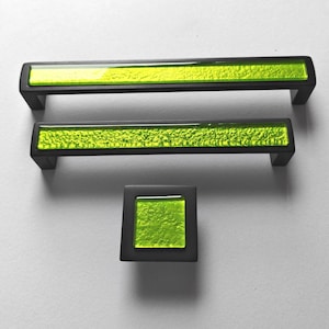 Op de afbeelding: Set van drie zwarte kastbeslagstukken: twee rechthoekige handgrepen en een vierkante knop. Elk stuk heeft een levendig, getextureerd, limoengroen midden in een zwart frame. De handgrepen zijn ongeveer 15 cm lang.