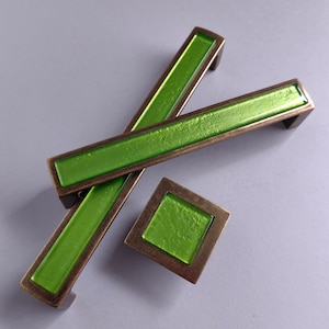 Op de afbeelding: Twee groene glazen kastgrepen met een bronzen afwerking. De grepen zijn rechthoekig en hebben een licht getextureerd oppervlak. Er is ook een kleinere vierkante greep met hetzelfde ontwerp.