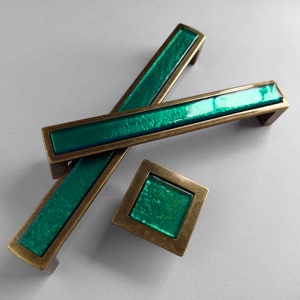 Może przedstawiać: Zestaw trzech zielonych szklanych i mosiężnych gałek do drzwi szafki. Gałki są prostokątne i mają fakturowaną powierzchnię.