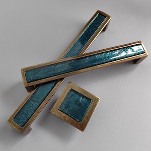May include: Two teal and brass cabinet pulls and a single teal and brass cabinet knob. The pulls are rectangular and the knob is square.