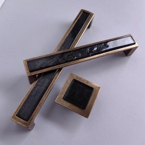May include: A set of three black and gold cabinet pulls with a rectangular and square shape. The pulls have a black, textured surface.