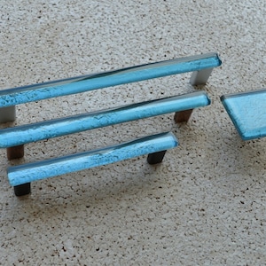 May include: A set of four cabinet hardware pieces. The set includes three rectangular pulls and one diamond-shaped knob. The hardware features a textured, light blue glass with white, brown, and black metal accents.