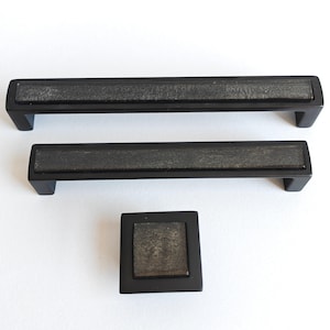 Peut inclure: Ensemble de trois pièces de quincaillerie d'armoire noires : deux poignées rectangulaires et un bouton carré. Chaque pièce a un centre texturé de couleur grise. Les poignées mesurent environ 20 cm de long.