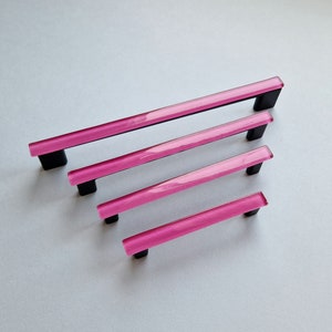 Peut inclure: Quatre poignées de porte de placard en verre rose avec des bases en métal noir. Les poignées sont disposées dans un ordre de taille décroissant.