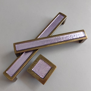 May include: Set of cabinet hardware featuring a rectangular and square design. The hardware has a bronze-colored frame with a textured, light purple center. The rectangular handles are approximately 6 inches long.