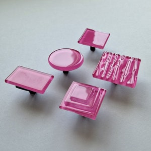 Peut inclure: Cinq boutons de cabinet en verre rose de différentes formes, notamment rondes, carrées et rectangulaires. Les boutons ont une apparence translucide et givrée.