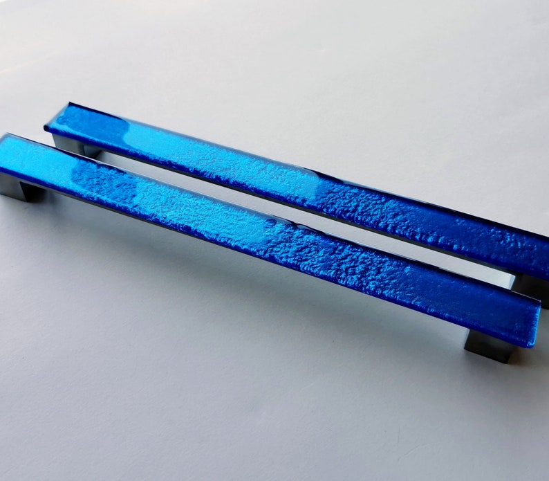 Set of two sparkly bright blue fired glass fridge pulls made of long glass sticks attached over a silvery metal base. Slightly textured glass furniture handles with rounded edges.