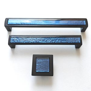 Könnte beinhalten: Set aus drei schwarzen Schrankbeschlägen. Zwei sind rechteckige Griffe mit einem strukturierten blauen Glaseinsatz, und einer ist ein quadratischer Knopf mit passendem blauem Glas. Für Schränke und Schubladen.