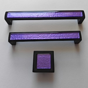 Peut inclure: Ensemble de trois pièces de quincaillerie d'armoire : deux poignées rectangulaires et un bouton carré. Les poignées et le bouton ont un cadre noir avec un centre violet texturé. Idéal pour les meubles et la décoration intérieure.