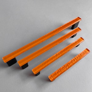May include: Four orange glass cabinet pulls with black metal mounting plates. The pulls are different sizes, with the largest pull at the top and the smallest pull at the bottom.