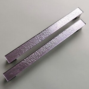 May include: Two silver cabinet pulls with a textured surface. Each pull is rectangular with a flat, smooth end. The pulls are angled slightly, showcasing their metallic sheen. These hardware pieces are designed for furniture.