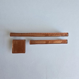 May include: A collection of copper-colored glass pieces. The set includes a square tile, a long rectangular bar, and a shorter rectangular bar. The glass has a reflective surface, and the pieces are arranged on a light gray background.