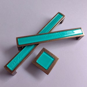 May include: Three teal blue glass cabinet knobs with a bronze finish. The knobs are in the shape of a rectangle and a square.