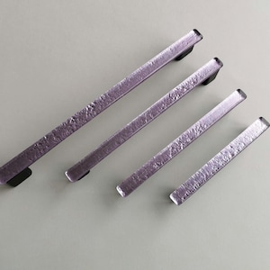 Könnte beinhalten: Vier violette Glas-Schranktürgriffe mit strukturierter Oberfläche. Die Griffe sind rechteckig und haben eine schwarze Metall-Montageplatte.