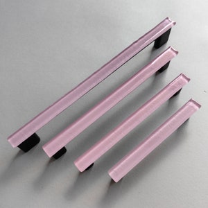 Peut inclure: Quatre poignées de tiroir en verre rose avec des plaques de montage en métal noir. Les poignées sont de tailles différentes, la plus grande en haut et la plus petite en bas.