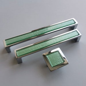 Könnte beinhalten: Drei Chrom-Schrankgriffe mit grünen Glaseinsätzen. Die Griffe sind in verschiedenen Größen, wobei der kleinste quadratisch ist.