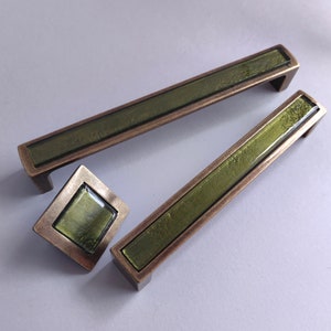 Op de afbeelding: Drie antieke messing kastgrepen met groene glazen inzetstukken. De grepen zijn rechthoekig en hebben een licht gebogen ontwerp. De glazen inzetstukken zijn diep groen en hebben een textuur.