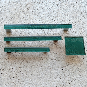 May include: A set of four teal glass cabinet hardware pieces. The set includes three rectangular pulls of varying lengths and a square knob. The hardware has a textured surface and is mounted on silver-toned metal bases.