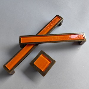 May include: Three orange glass cabinet knobs with a brass finish. The knobs are in the shape of a rectangle and a square.