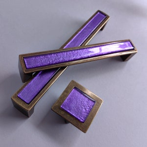 May include: Three purple glass cabinet knobs with a bronze metal frame. The knobs are in the shape of a rectangle and a square.