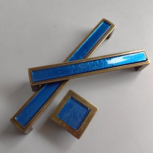 May include: Three blue glass cabinet knobs with a brass finish. The knobs are rectangular and square in shape.