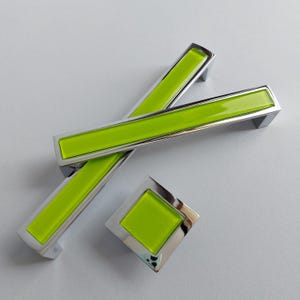 Puede incluir: Un juego de herrajes para armarios con un inserto rectangular verde brillante dentro de un marco plateado pulido. El juego incluye dos tiradores largos y rectangulares y un pomo cuadrado, todos con un diseño moderno.